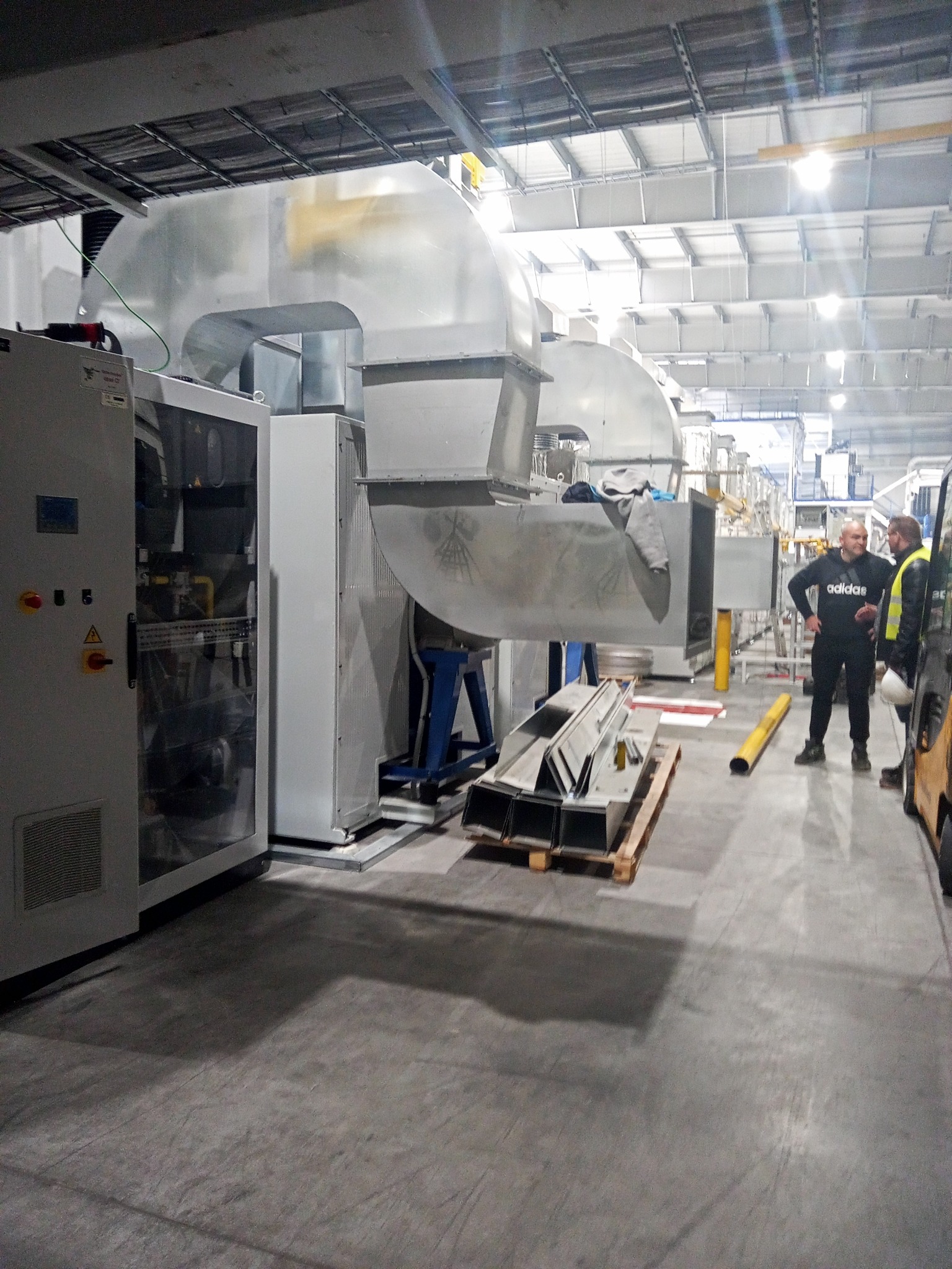 Wnętrze hali produkcyjnej z systemem wentylacyjnym w trakcie instalacji, widoczne kanały wentylacyjne, szafa sterownicza i paleta z elementami montażowymi, dwóch mężczyzn w tle.