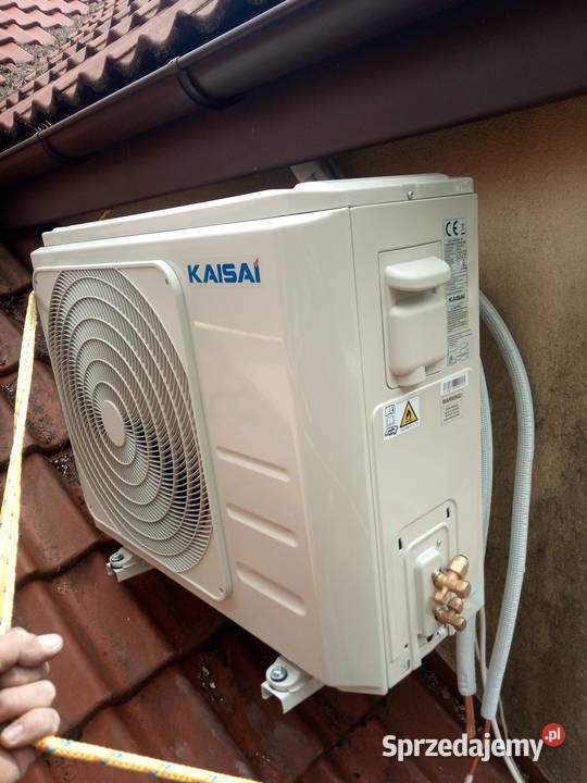 Boczny widok jednostki zewnętrznej klimatyzacji Kaisai zamontowanej na dachu z czerwonej dachówki, obok rynny. Widoczne przyłącza i przewody. W kadrze fragment dłoni trzymającej linę.