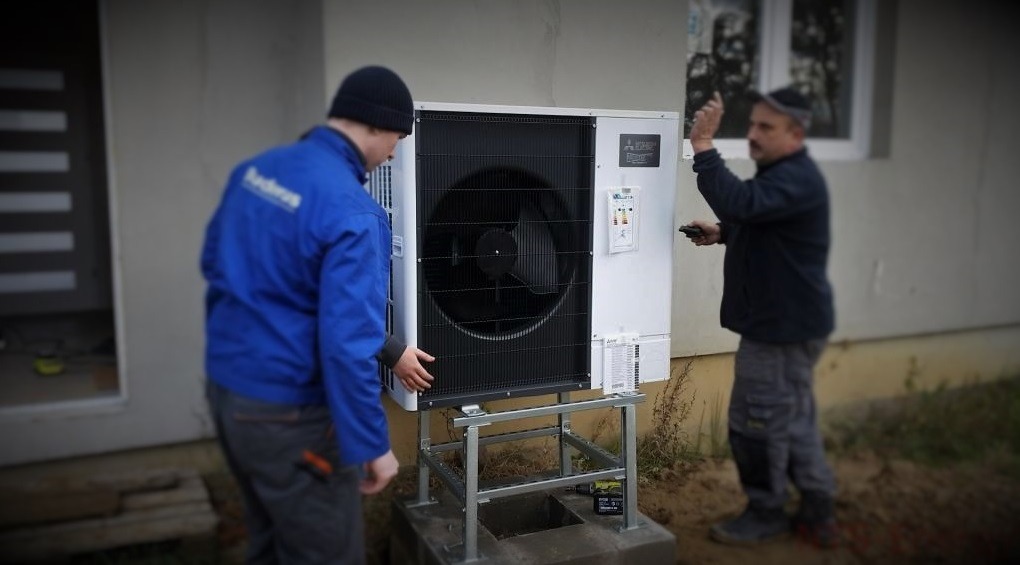 Dwóch instalatorów montuje zewnętrzną jednostkę grzewczą na metalowej konstrukcji przed budynkiem z szarą elewacją. Jeden instalator w niebieskiej kurtce dotyka obudowy, drugi trzyma pilot.