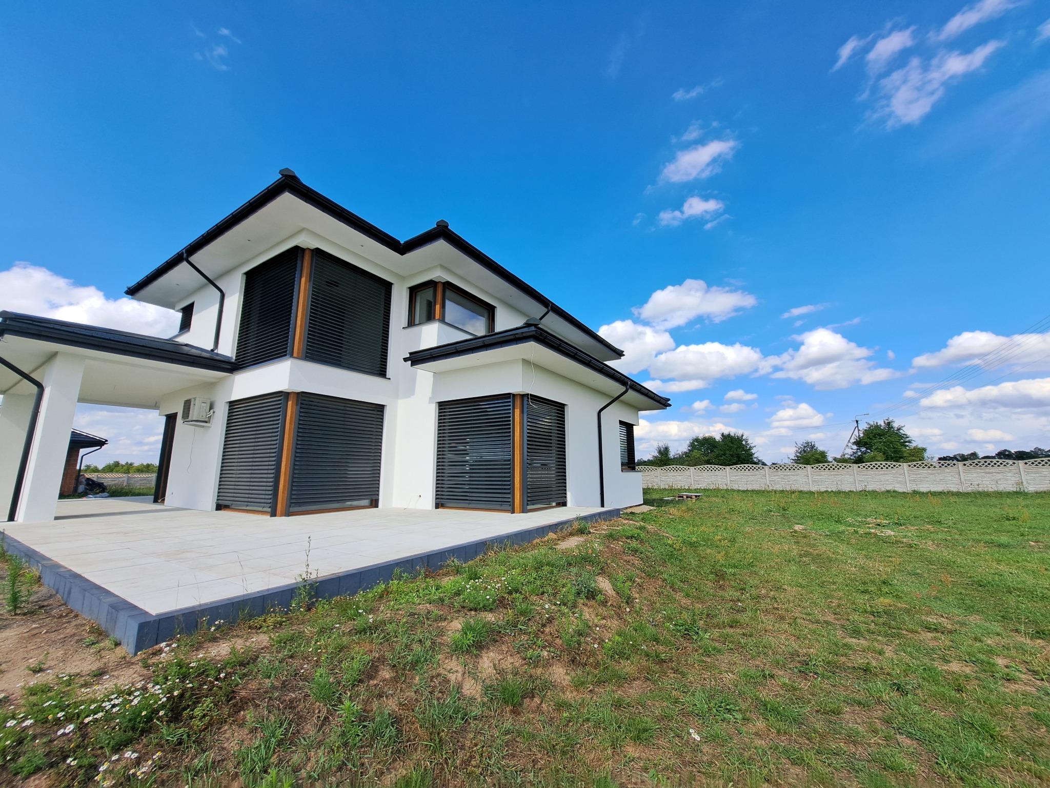 Nowoczesny, biały dom z płaskim dachem i ciemnymi żaluzjami zewnętrznymi, widziany z poziomu gruntu na tle błękitnego nieba z obłokami.