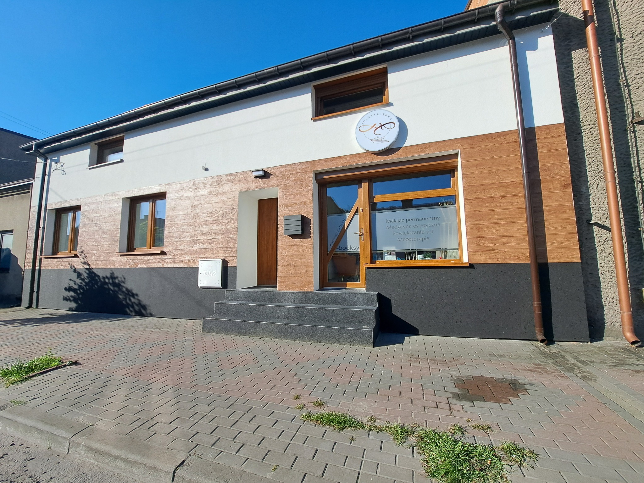 Dwukondygnacyjny budynek z elewacją w kolorach bieli, brązu i grafitu, z oknami w drewnianych ramach i szyldem reklamowym gabinetu kosmetycznego Joanna Lakoma.
