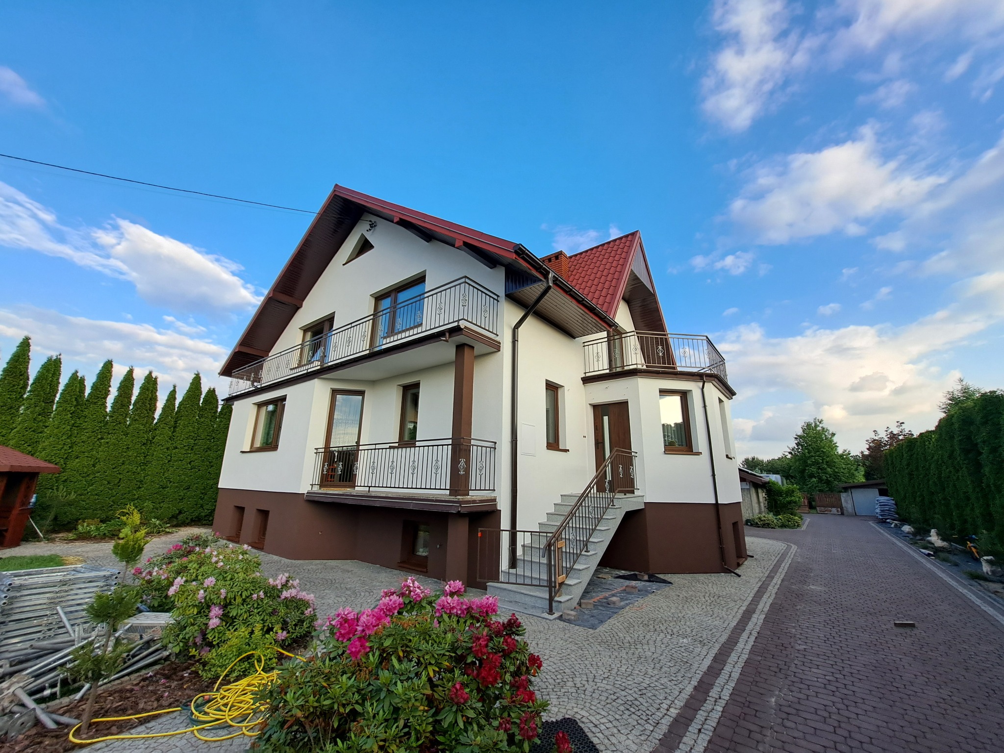 Dwupiętrowy dom z wykończoną elewacją w kolorze białym i brązowym, z balkonami z metalowymi balustradami, czerwonym dachem i schodami wejściowymi, otoczony zielenią i brukowaną ścieżką.