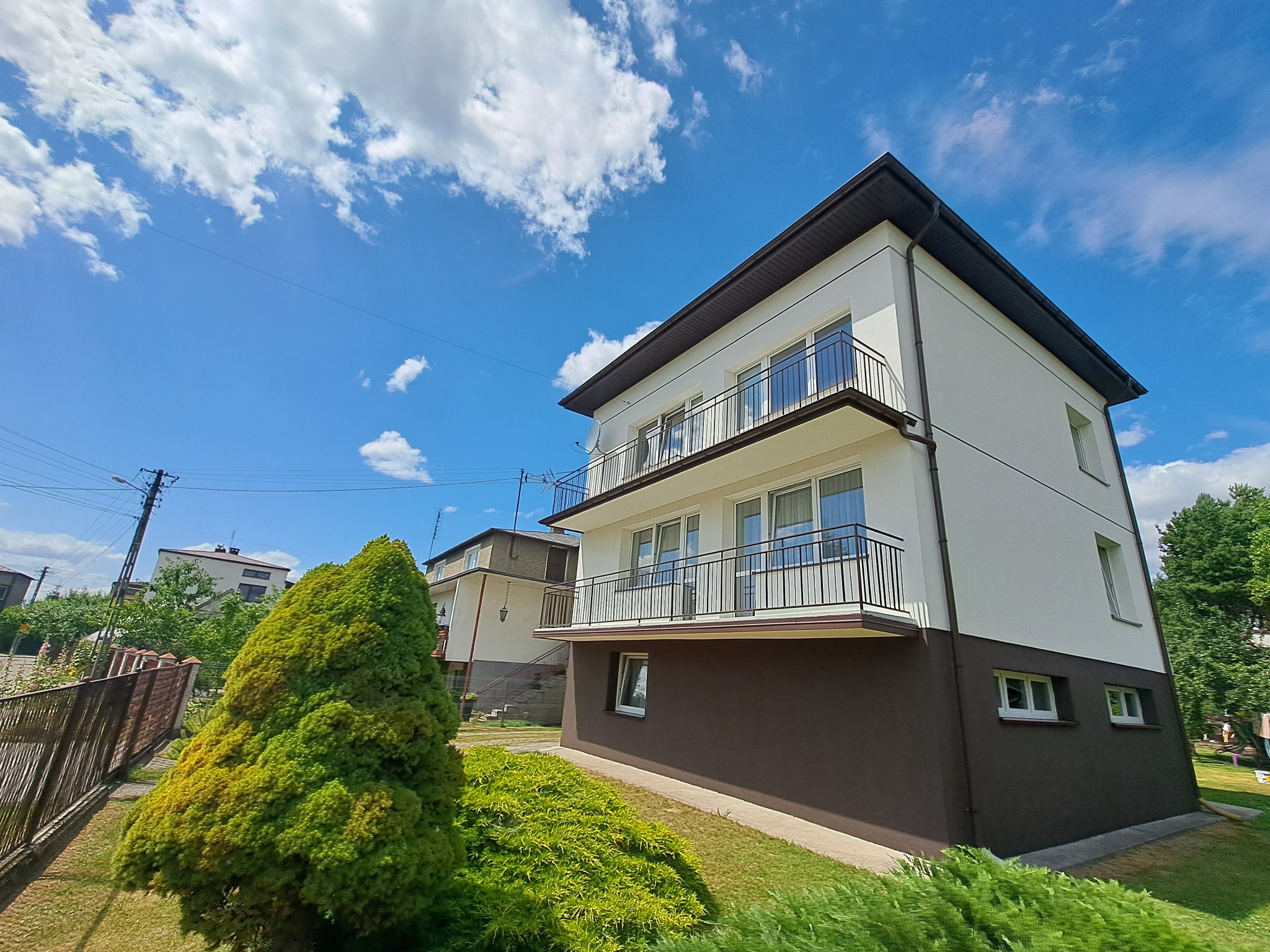 Dwukondygnacyjny dom z balkonami, świeżo po malowaniu elewacji w kolorach bieli i brązu, na tle błękitnego nieba z chmurami. Widok z dołu.