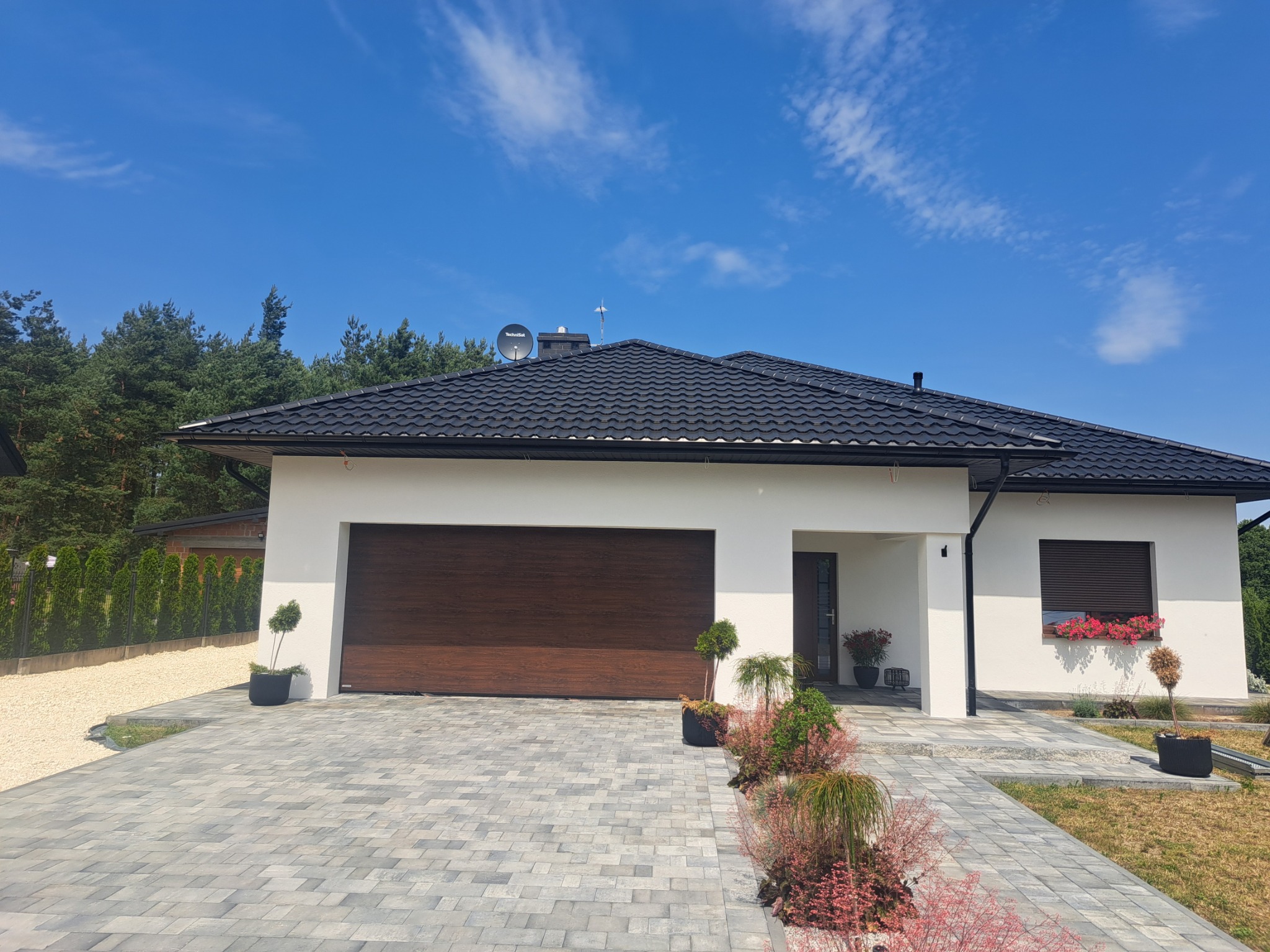 Nowoczesny dom jednorodzinny z białą elewacją, brązową bramą garażową i elegancką kostką brukową na podjeździe, w słoneczny dzień.