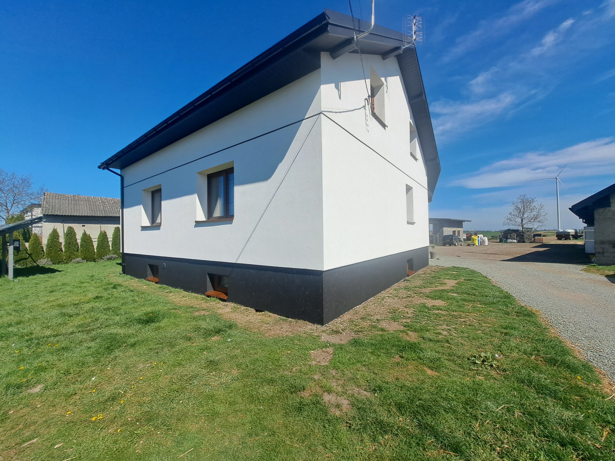 Biały dom z ciemną podmurówką i dachem na tle błękitnego nieba. Widoczna antena na dachu, trawnik i droga. W tle turbina wiatrowa.