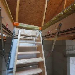 Drewniane schody strychowe, składane, z metalowymi sprężynami, zamontowane w ocieplonym poddaszu z widocznymi elementami konstrukcyjnymi i bloczkami betonowymi.