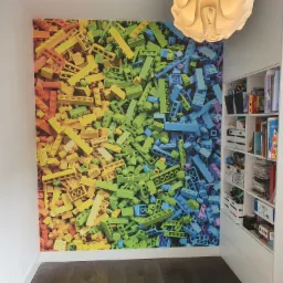 Ściana w pokoju dziecięcym z fototapetą przedstawiającą klocki w układzie kolorystycznym tęczy, obok biały regał z półkami wypełnionymi książkami i zabawkami, wisząca lampa w kształcie kwiatu.