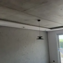 Wnętrze w trakcie wykańczania: szara ściana z efektem dekoracyjnym, surowy betonowy sufit z oświetleniem LED i wisząca lampa w stylu retro, okna z planami architektonicznymi przyklejonymi taśmą.