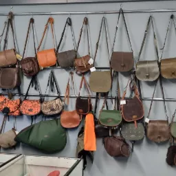 Ekspozycja różnorodnych toreb skórzanych i materiałowych w sklepie, wiszących na metalowych relingach na szarej ścianie, obok regału z innymi wyrobami galanteryjnymi i siatki z akcesoriami.