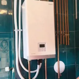 Biały, wiszący piec grzewczy marki Viessmann z podłączonymi miedzianymi rurami i białymi przewodami, zamontowany na ścianie wyłożonej zielonymi płytkami.