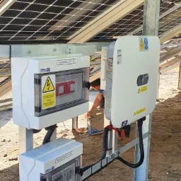 Rozdzielnice PV-DC i PV-AC oraz falownik zamontowane pod panelami słonecznymi, widoczne kable i ziemiste podłoże.