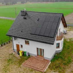 Dom jednorodzinny z ciemną dachówką i panelami słonecznymi na dachu, widok z góry, na trawniku stoją trzy pojemniki na śmieci.