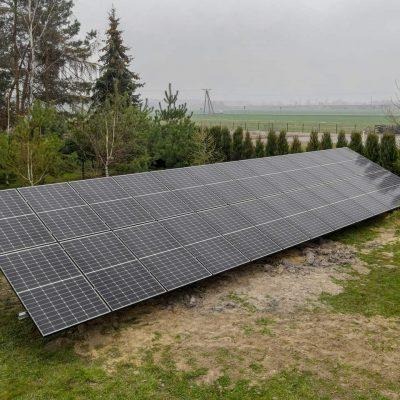 Instalacja fotowoltaiczna na gruncie, widoczne panele w układzie poziomym, w tle drzewa i pole w pochmurny dzień.