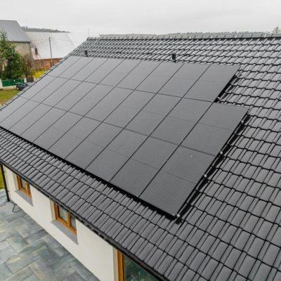 Czarny panel słoneczny na dachu domu z ciemnej dachówki, widok z góry, pochmurne niebo w tle.