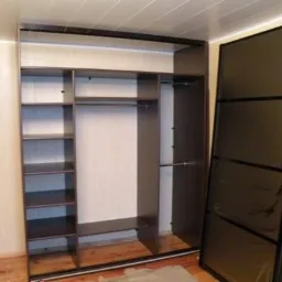 Wnętrze szafy przesuwnej z półkami i drążkami, z odstawionymi drzwiami w kolorze ciemnego brązu.