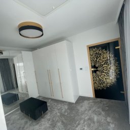 Solidne sprzątanie - remonty - Wnętrze pokoju po remoncie: białe, błyszczące szafy w zabudowie z podłużnymi, złotymi uchwytami, czarny puf z weluru na złotych nóżkach, drzwi z czarnym skrzydłem i złotym, roślinnym ornamentem...