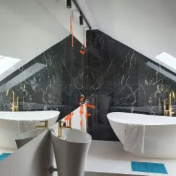 Nowoczesna łazienka na poddaszu z czarnymi marmurowymi płytkami, wolnostojącą białą wanną, złotą armaturą i minimalistyczną umywalką, częściowo zabezpieczona pomarańczową folią ochronną.