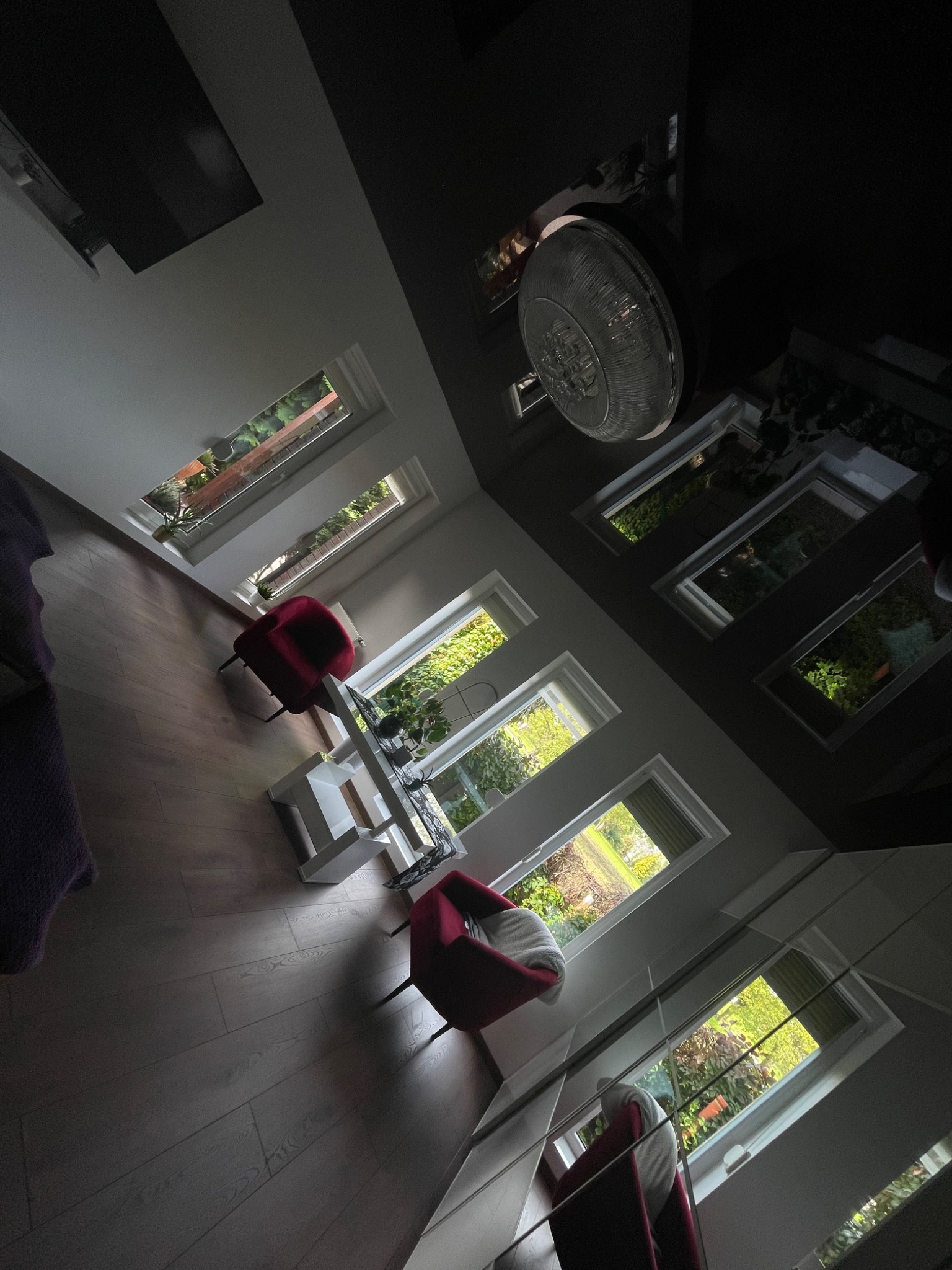 Nowoczesne wnętrze z parkietem, czerwonymi fotelami i oknami dachowymi z widokiem na zieleń, oświetlone centralnym, okrągłym żyrandolem.