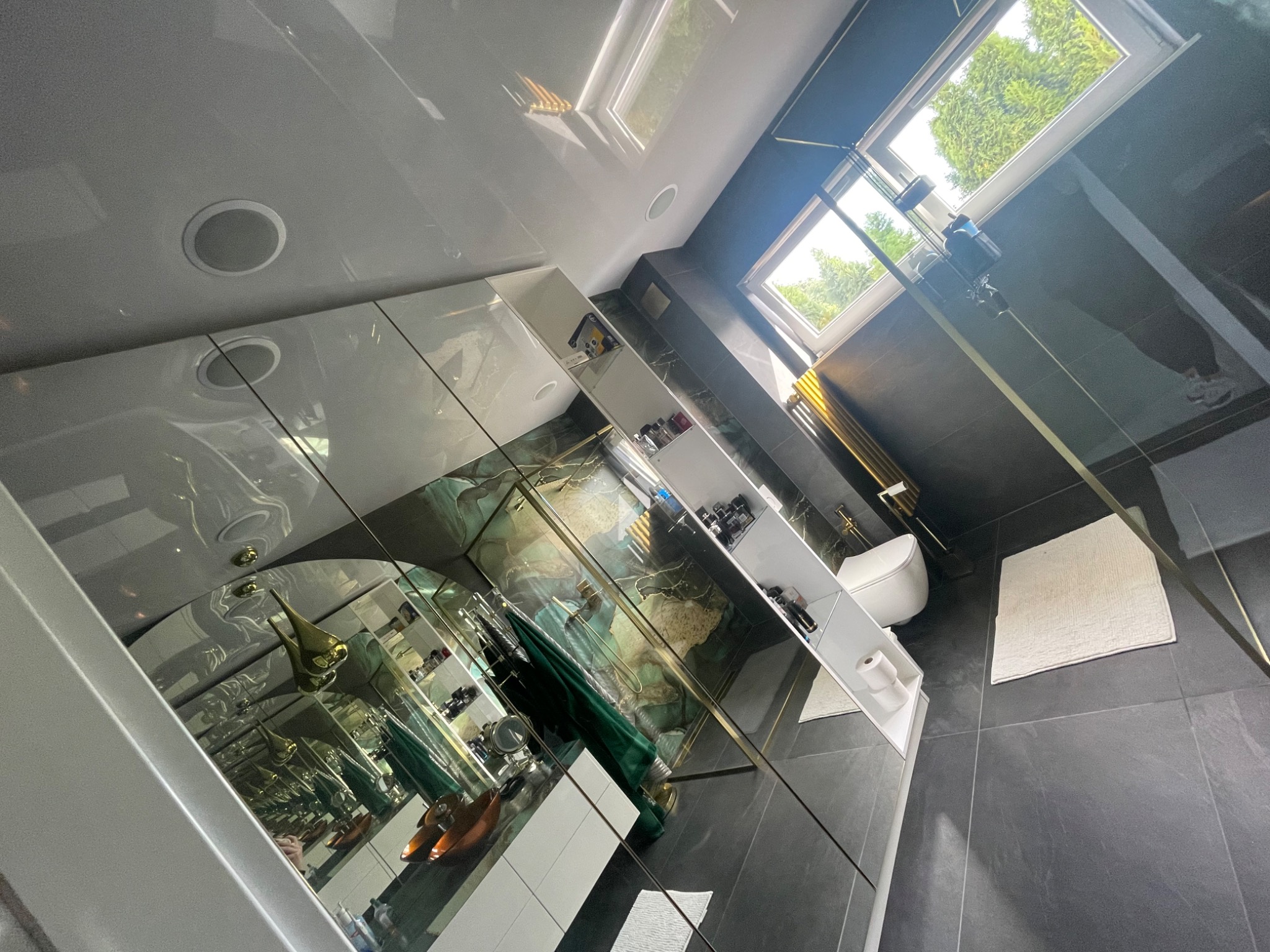 Nowoczesna łazienka z czarnymi płytkami, szklaną kabiną prysznicową ze złotymi elementami, wiszącą toaletą i oknem dachowym wpuszczającym naturalne światło.