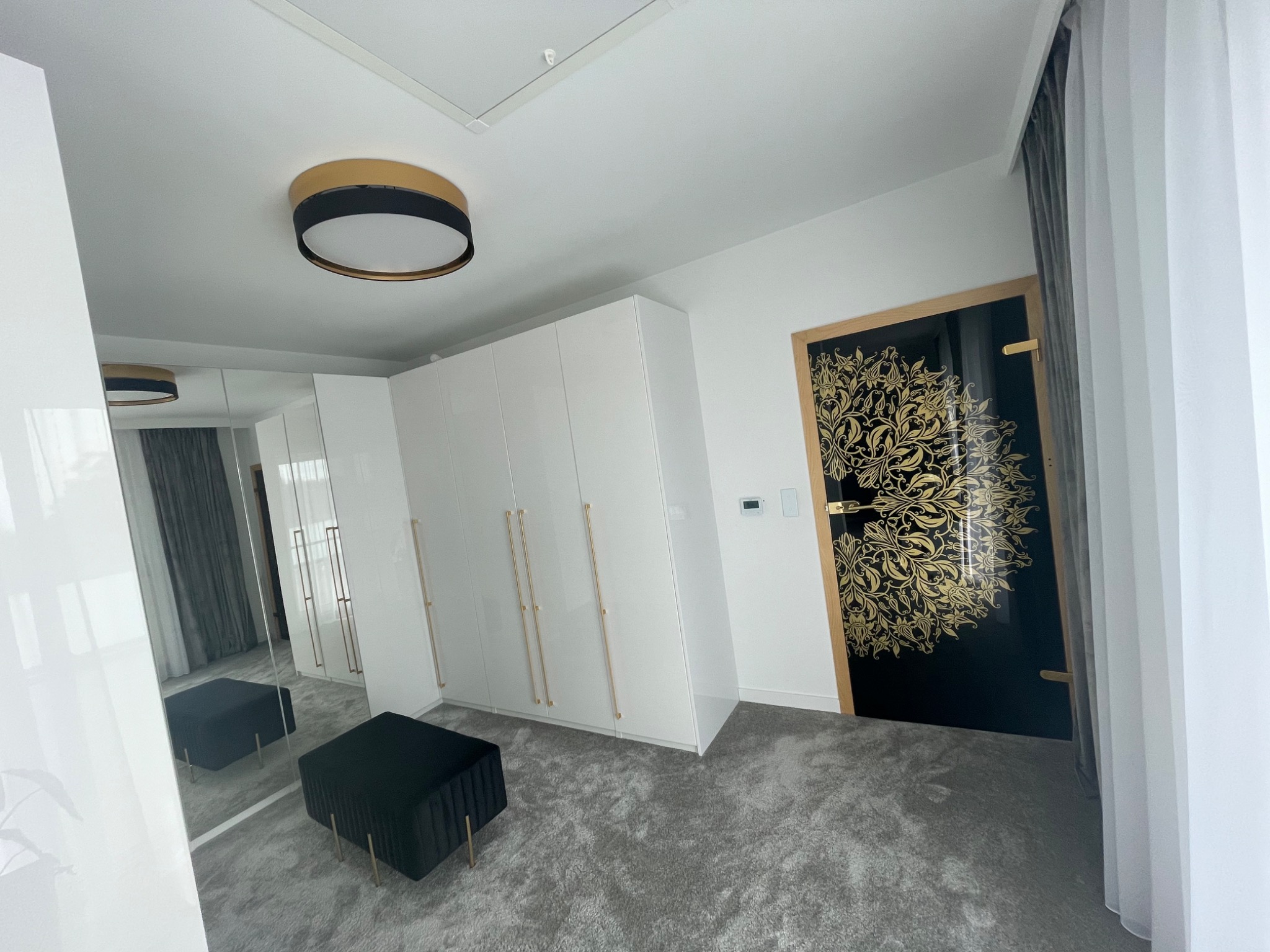 Wnętrze pokoju po remoncie: białe, błyszczące szafy w zabudowie z podłużnymi, złotymi uchwytami, czarny puf z weluru na złotych nóżkach, drzwi z czarnym skrzydłem i złotym, roślinnym ornamentem...