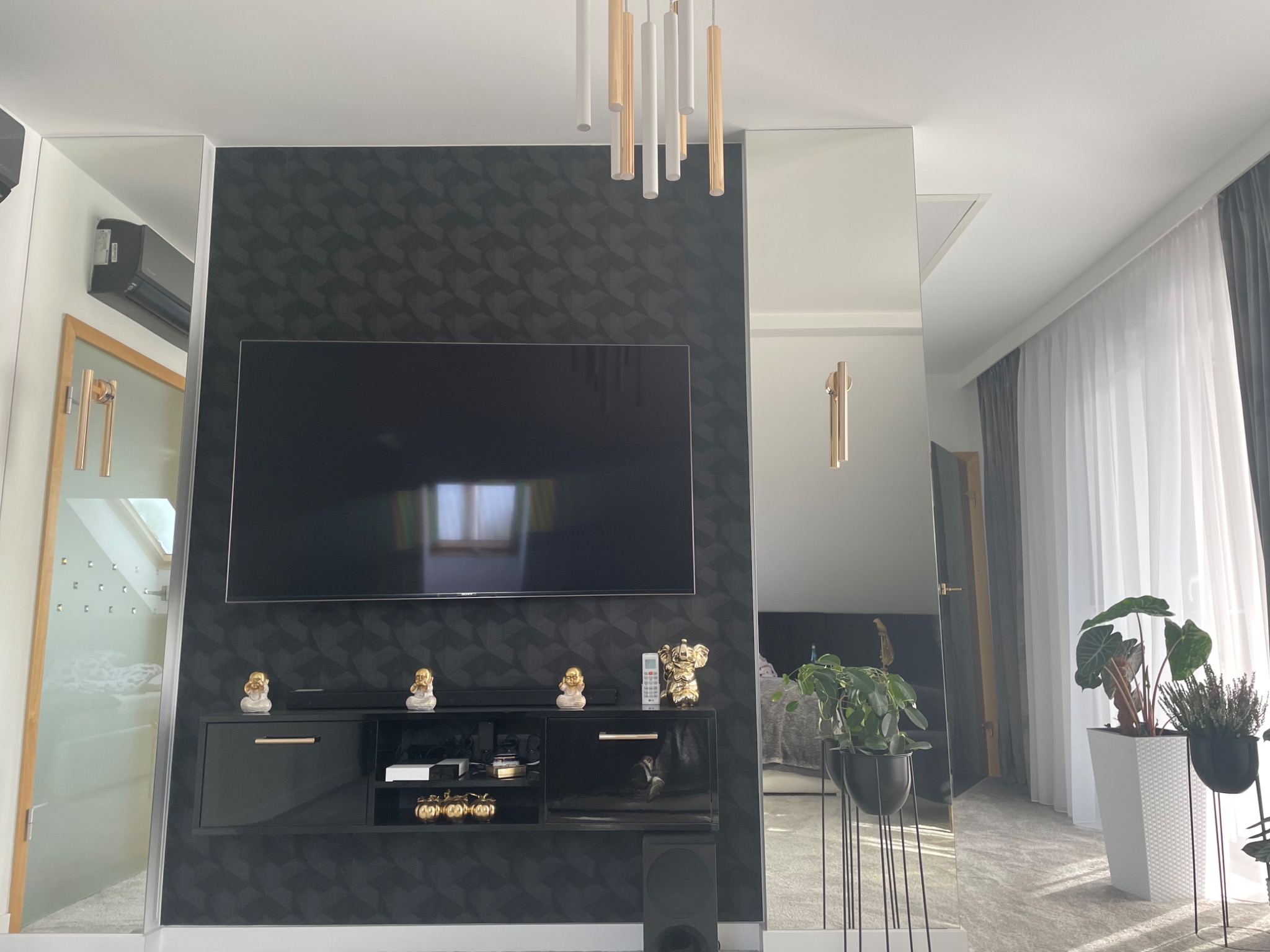 Nowoczesny salon z czarną ścianą akcentową o geometrycznym wzorze, telewizorem, błyszczącą czarną szafką RTV z dekoracyjnymi figurkami złotych Buddów i słonia, oraz lustrami po bokach, odbijającymi...