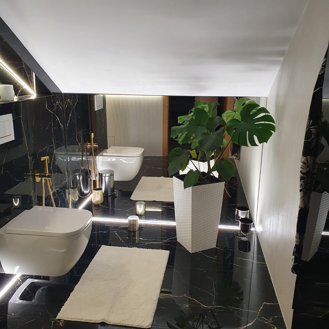 Elegancka łazienka z czarnymi marmurowymi płytkami, podwieszanymi toaletami, złotymi akcentami armatury i dekoracyjnymi świecami, oświetleniem LED pod sufitem i dużą rośliną Monstera w białej donicy.
