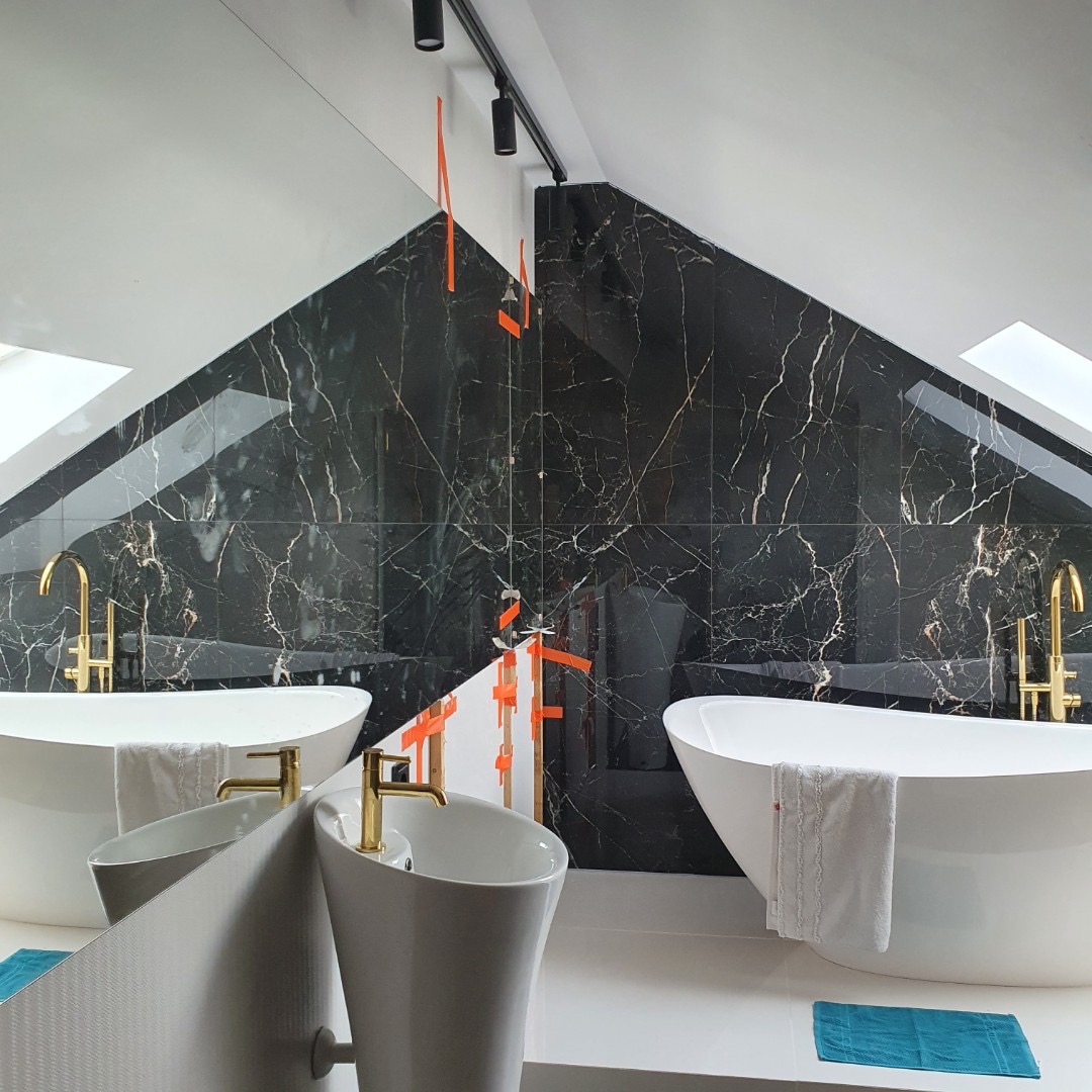 Nowoczesna łazienka na poddaszu z czarnymi marmurowymi płytkami, wolnostojącą białą wanną, złotą armaturą i minimalistyczną umywalką, częściowo zabezpieczona pomarańczową folią ochronną.