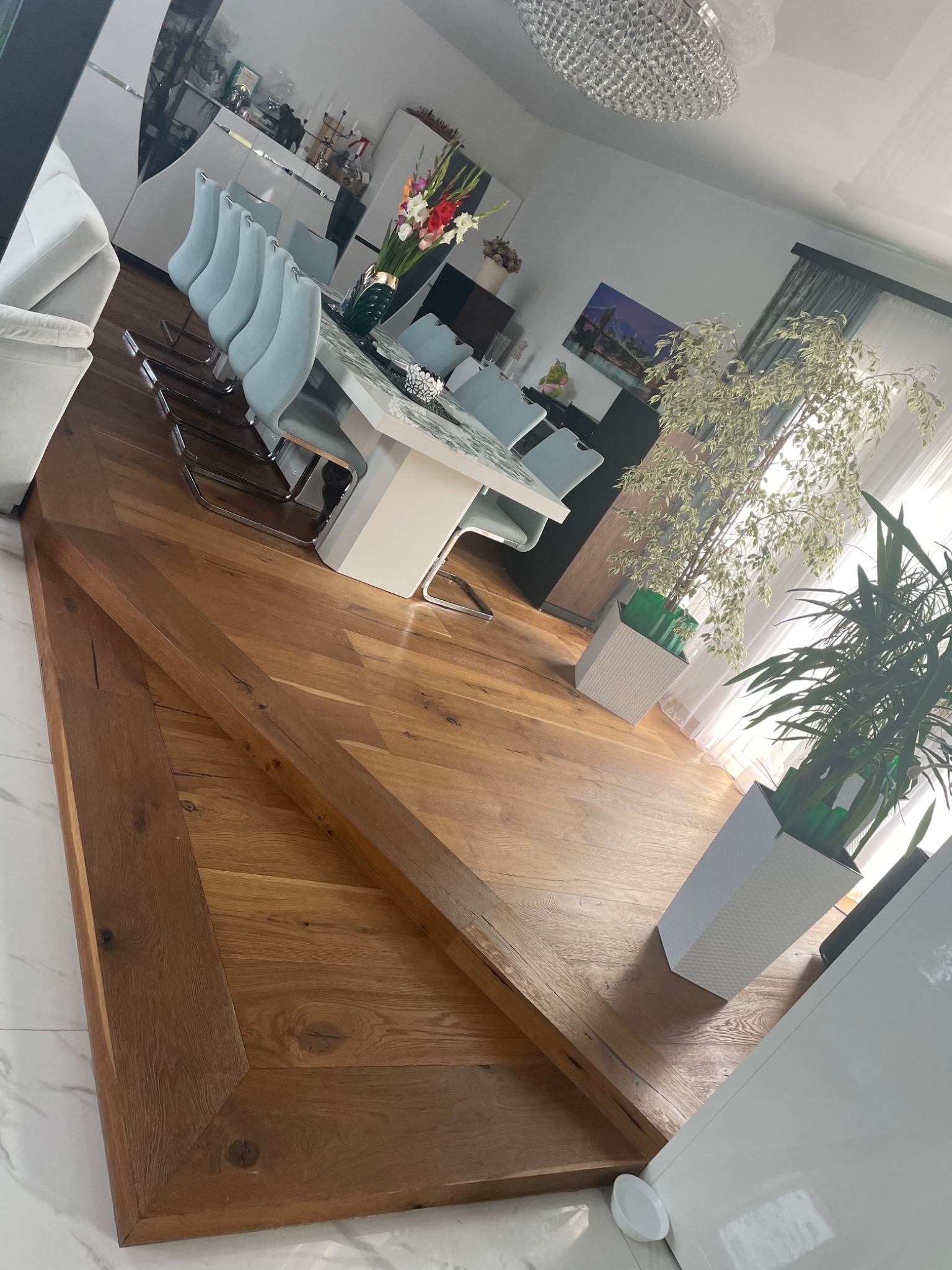 Nowoczesna jadalnia z drewnianą podłogą w jodełkę, białym stołem i krzesłami w odcieniach błękitu, doświetlona wiszącym kryształowym żyrandolem i naturalnym światłem wpadającym przez okno.