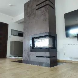 Nowoczesny kominek obudowany szarobrązową płytą imitującą beton, z czarnymi akcentami w postaci obramowania paleniska i poziomych wnęk, w jasnym salonie z drewnianą podłogą.