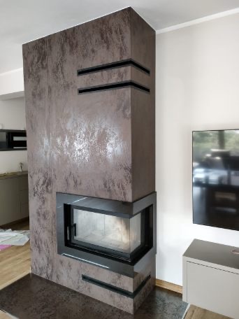 Nowoczesny kominek narożny z obudową w odcieniach brązu i czarnymi akcentami, wkomponowany w przestrzeń salonu z telewizorem w tle.