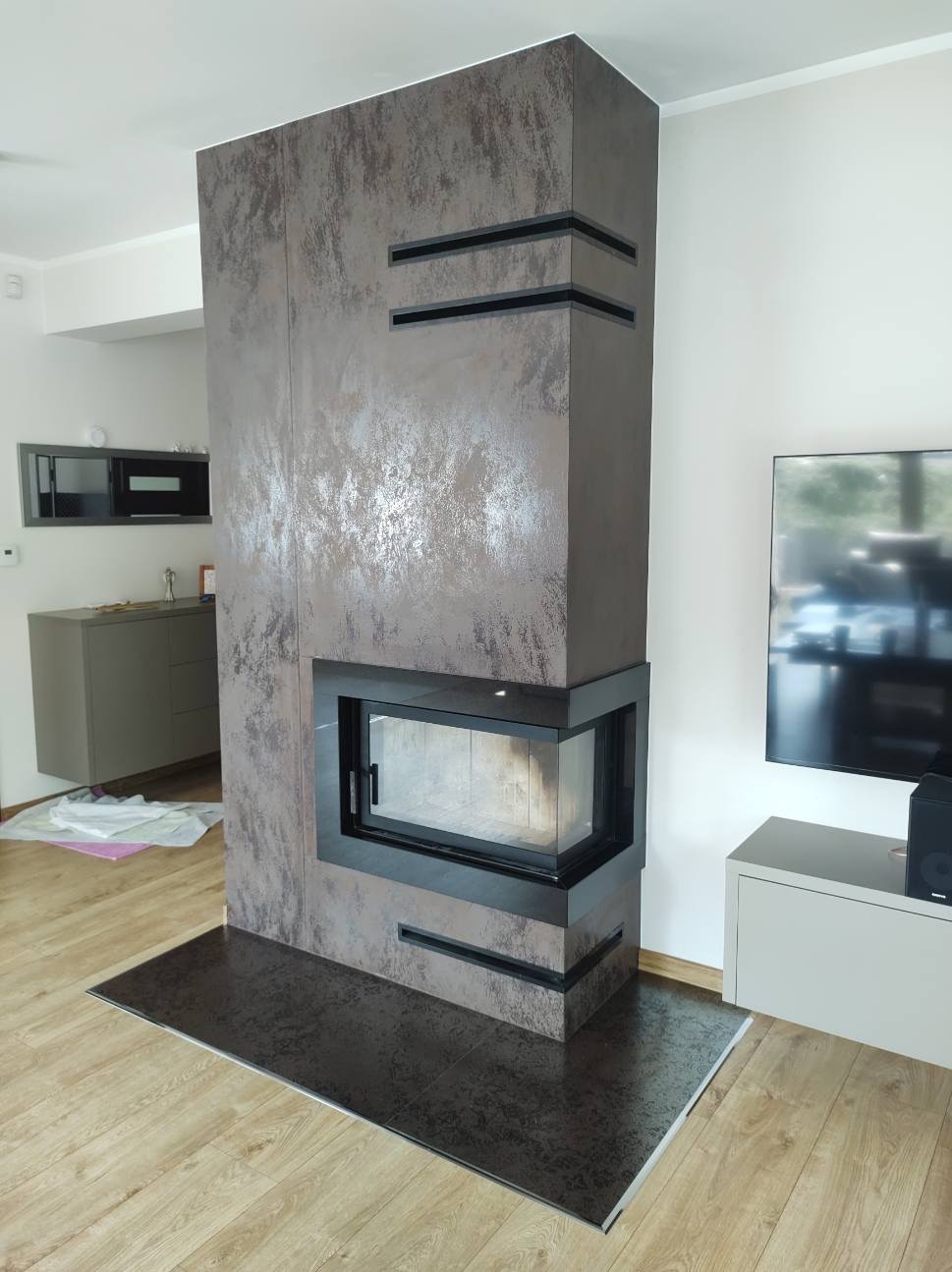 Nowoczesny kominek narożny z obudową w odcieniach szarości i brązu, z czarnymi akcentami, na tle jasnego salonu z telewizorem.