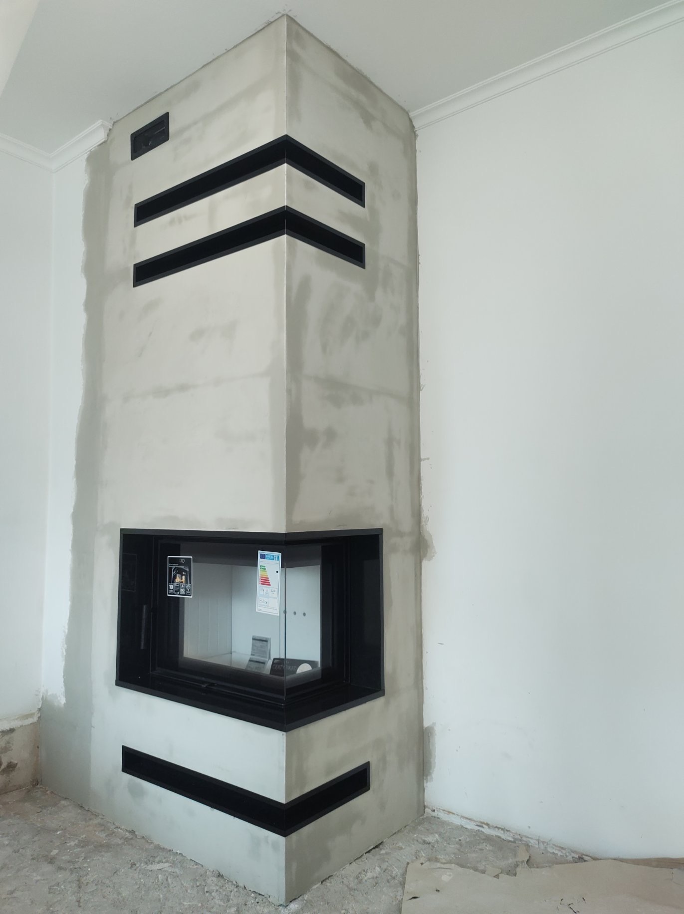 Narożny kominek z czarnymi, poziomymi wstawkami w surowym wykończeniu, widoczne niedoskonałości tynku i resztki zaprawy na podłodze.