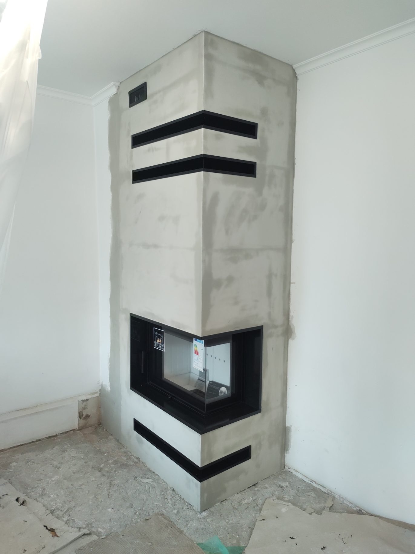 Narożny kominek w surowej obudowie z betonu architektonicznego, z czarnymi, poziomymi wstawkami, w trakcie montażu, widoczne niedokończone ściany i podłoga.