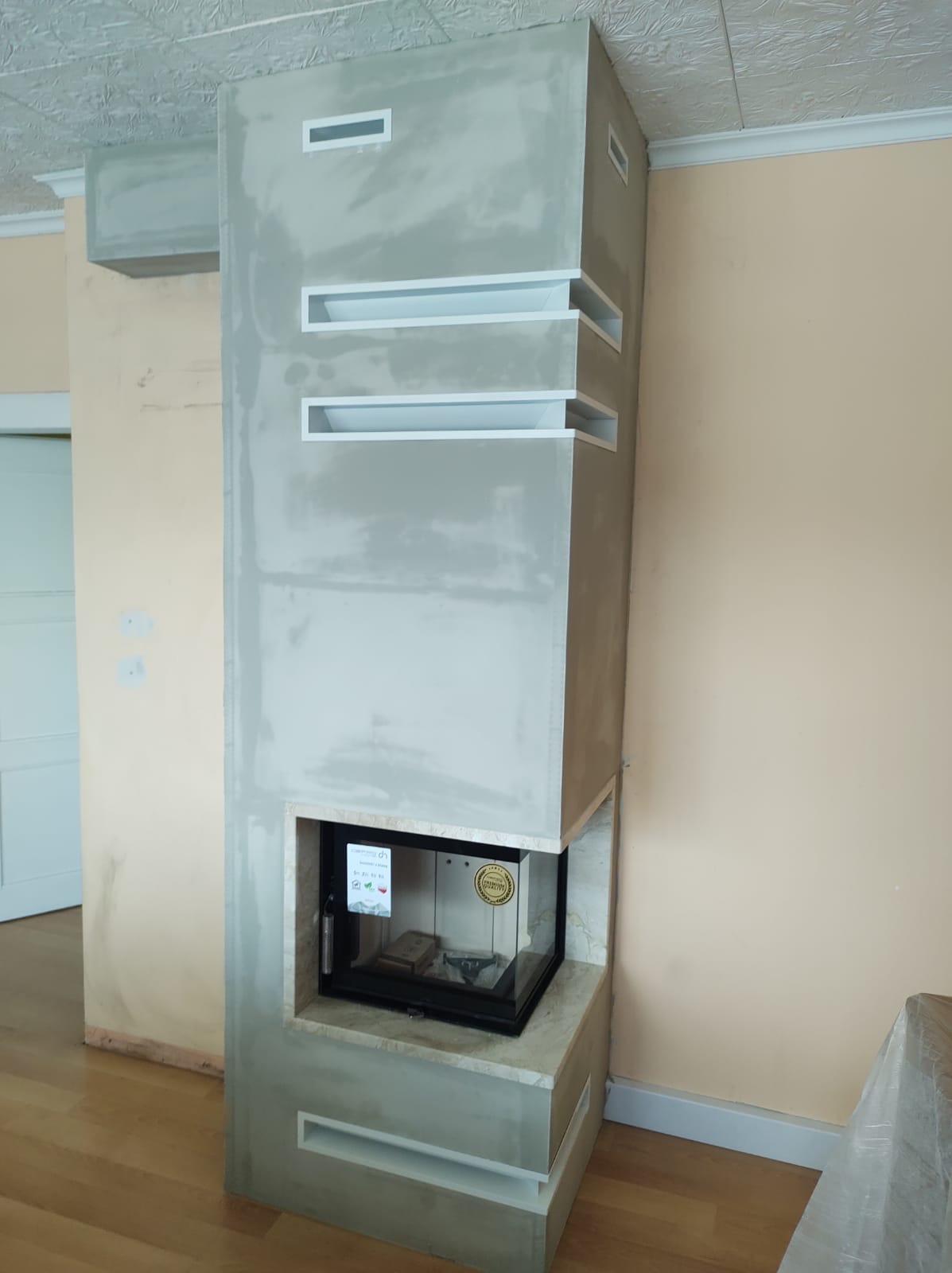 Nowoczesna obudowa kominkowa w trakcie wykańczania, surowy tynk w odcieniach szarości, z wnękami wykończonymi na biało, widoczna szyba kominka.