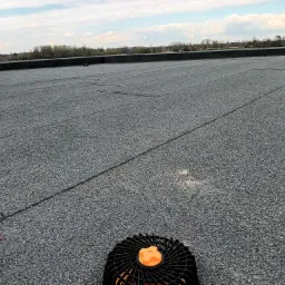 Szary, płaski dach pokryty papą z widocznymi zgrzewami, na pierwszym planie odpowietrznik dachowy, w tle zabudowania przemysłowe i zieleń.