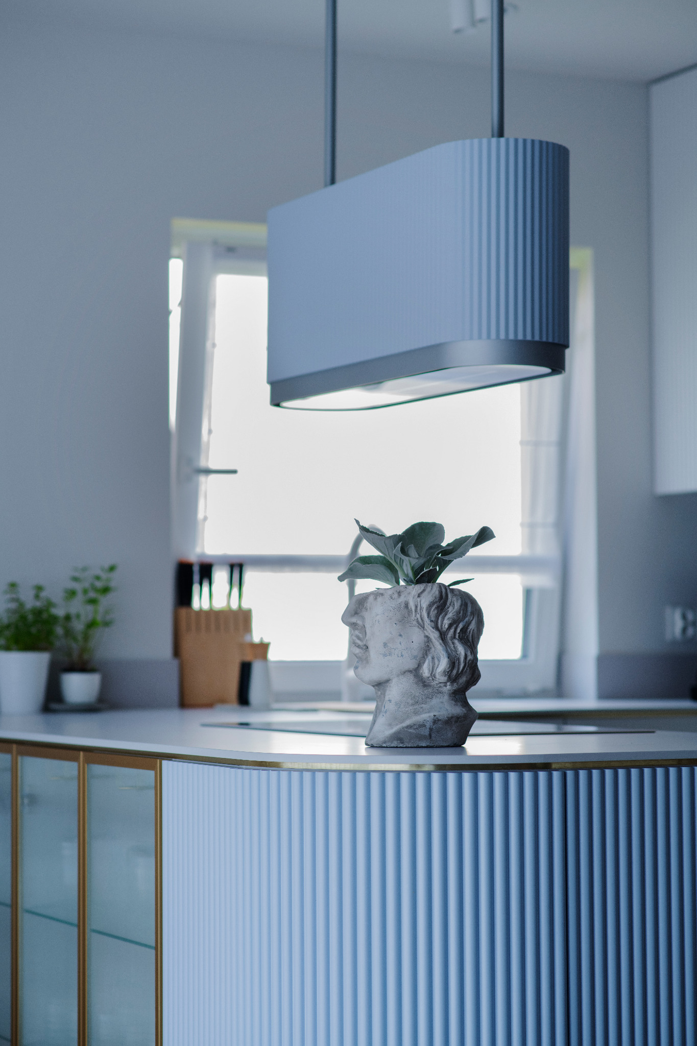 Niebieska, prążkowana wyspa kuchenna z blatem i złotymi akcentami. Na blacie rzeźba głowy z rośliną. W tle okno i lampa. Minimalistyczny wystrój kuchni.