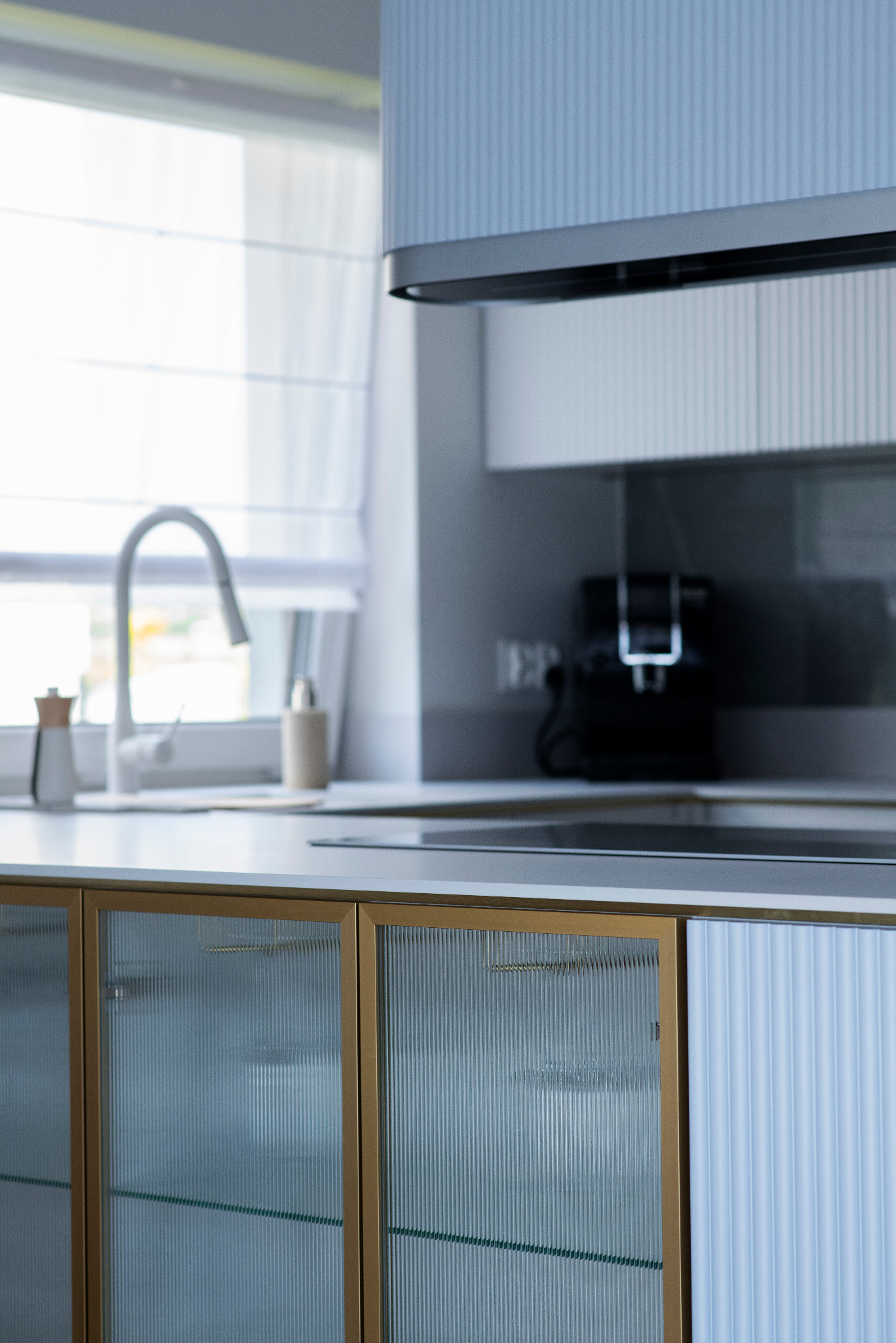 Nowoczesna kuchnia z ryflowanymi frontami szafek w odcieniach błękitu i złota. Widoczne szklane drzwi szafek, bateria kuchenna i ekspres do kawy w tle.