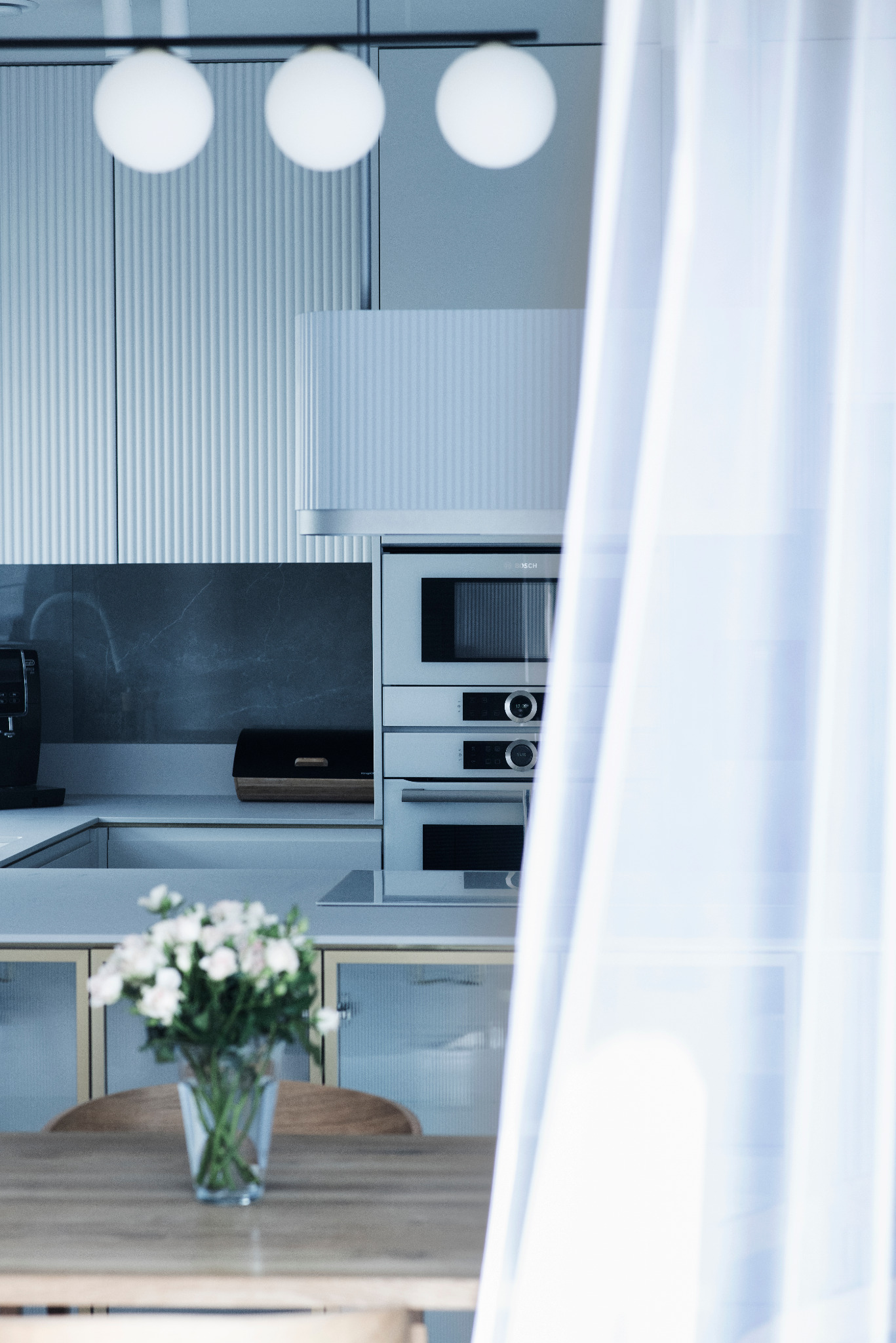 Jasna kuchnia z frezowanymi frontami szafek, zabudowanym sprzętem AGD i blatem z szarego kamienia, uzupełniona drewnianym stołem i bukietem białych kwiatów.