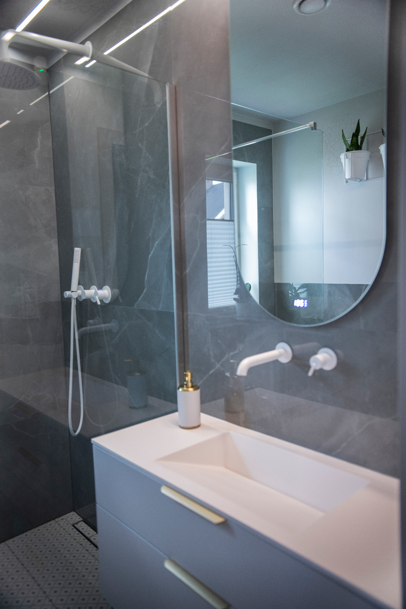 Nowoczesna łazienka z szarą armaturą i umywalką wpuszczaną w szafkę z złotymi uchwytami. W tle kabina prysznicowa i okrągłe lustro z wyświetlaczem.