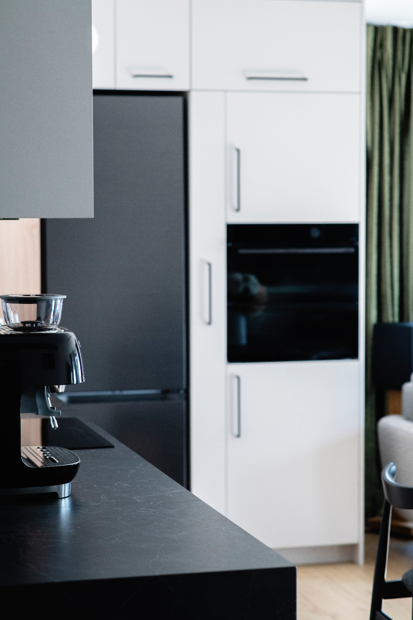 Nowoczesna kuchnia z czarnym blatem i ekspresem do kawy. Białe szafki, czarna lodówka i piekarnik w zabudowie. Minimalistyczny design z zielonymi zasłonami w tle.