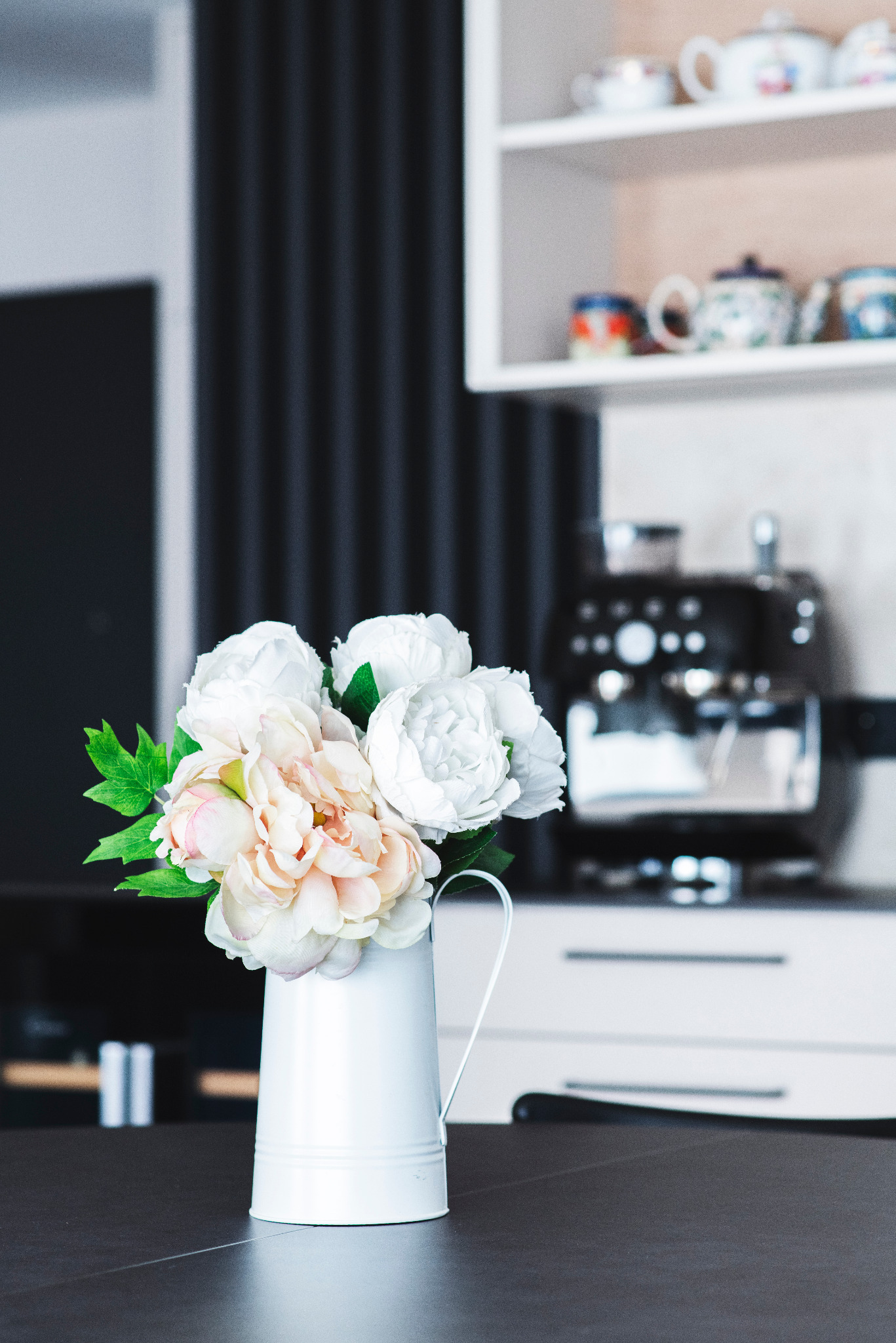 Dekoracyjny bukiet w białym dzbanku na tle nowoczesnej kuchni z ekspresem do kawy i półkami z kolekcją czajników.