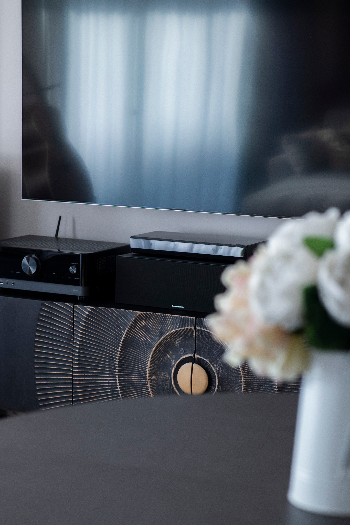 Czarna szafka RTV z dekoracyjnym frontem, na której stoi sprzęt audio. W tle telewizor i bukiet kwiatów. Ujęcie z bliska, podkreślające detale wykończenia.