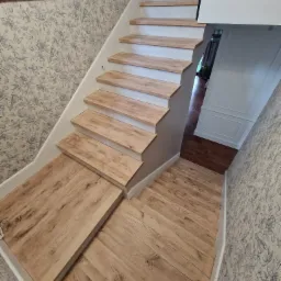 Jasne schody z drewnianymi stopniami i białymi podstopnicami, widok z góry na tle ściany w kwiatowy wzór.