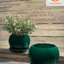 Dwie ciemnozielone, ceramiczne donice kuliste na podstawkach, jedna z kwitnącą rośliną, logo producenta w prawym górnym rogu, na tle drewnianej ściany.