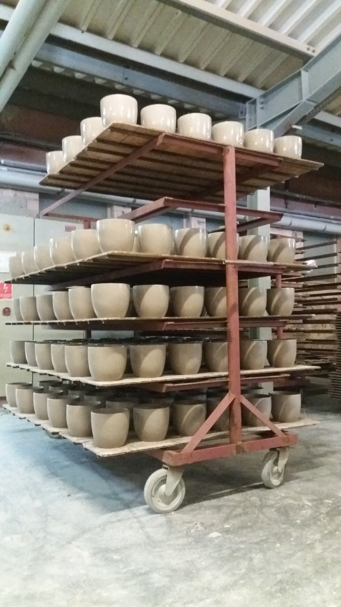 Wielopoziomowy wózek z metalową ramą wypełniony rzędami ceramicznych doniczek w jasnym odcieniu beżu, ustawionych w regularnych odstępach w hali produkcyjnej.