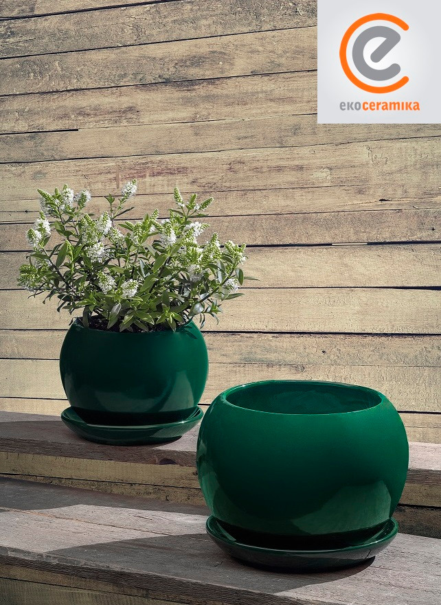 Dwie ciemnozielone, ceramiczne donice kuliste na podstawkach, jedna z kwitnącą rośliną, logo producenta w prawym górnym rogu, na tle drewnianej ściany.