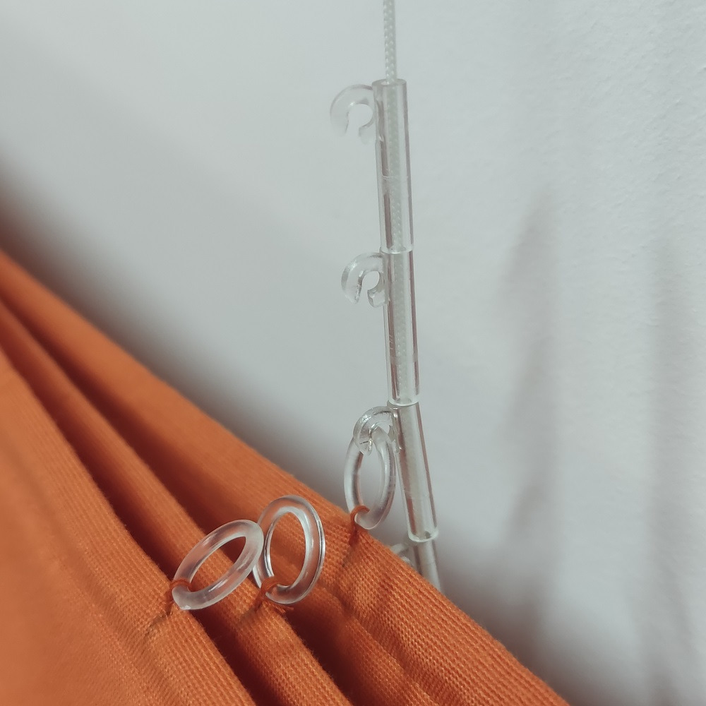 Pomarańczowa zasłona z przezroczystymi plastikowymi kółkami i systemem mocowania do karnisza, widoczne detale montażu.