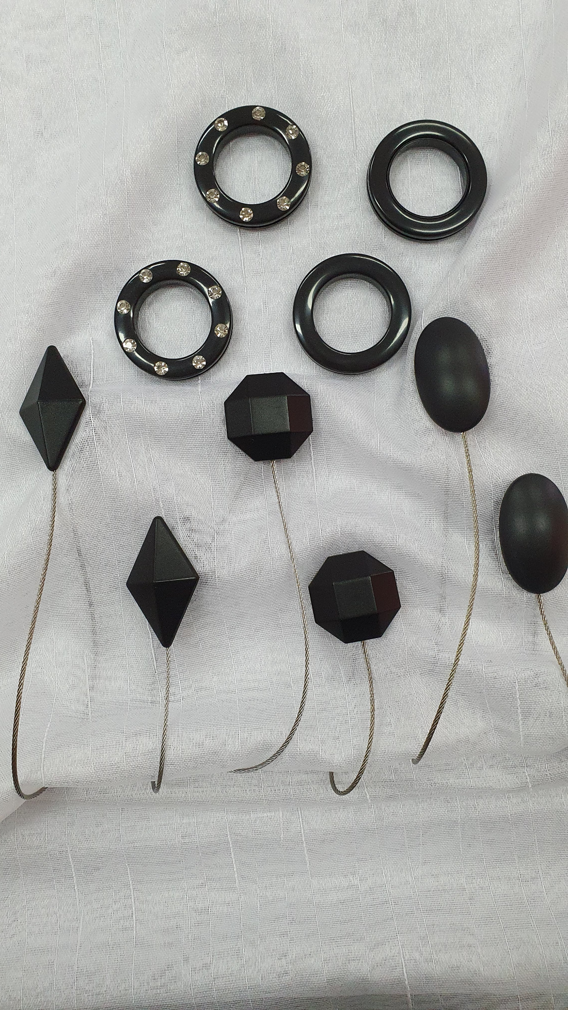 Zestaw czarnych kółek przelotowych do zasłon, niektóre z kryształkami, oraz haczyków do upinania zasłon o geometrycznych kształtach na białym, teksturowanym tle.