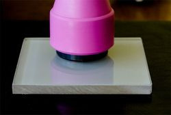 Zbliżenie na element dociskający do przezroczystej, kwadratowej płyty, prawdopodobnie podczas procesu produkcyjnego lub naprawczego.