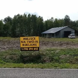 Żółty baner reklamowy z napisem 'Miejsce na Twoją Reklamę' i numerem telefonu, umieszczony na tle przygotowanego terenu z budynkiem magazynowym w oddali i lasem w tle.